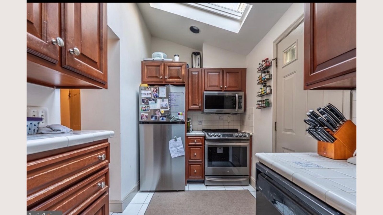 Existing narrow galley kitchen with dated cherry cabinetry, a wall microwave and tile counters